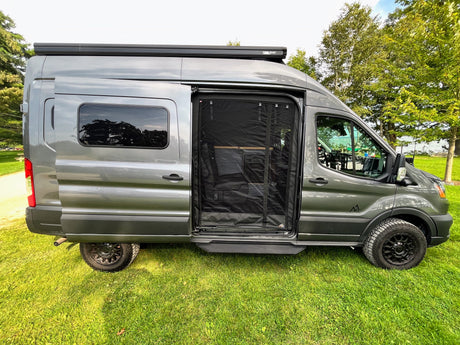 A sleek grey conversion van, set up as an RV rental, parked on grass with its side screen door open for fresh air.