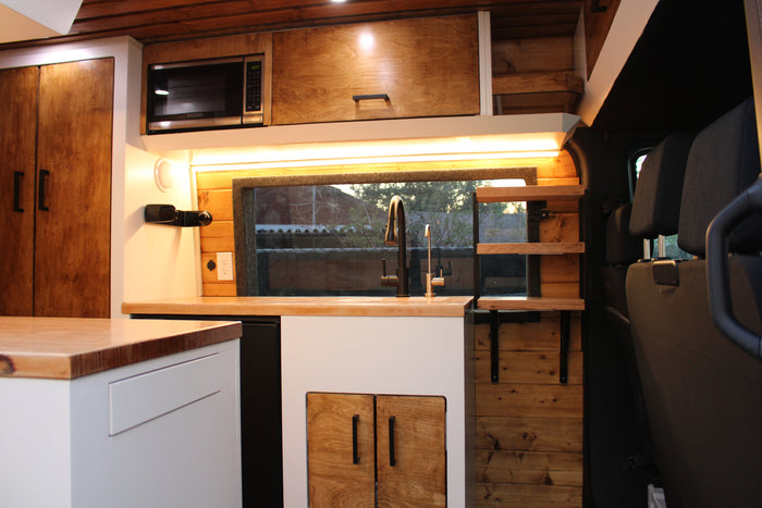 Cozy RV rental kitchen: wood cabinets, light counters, sink, microwave, and ambient lighting in a sleek van conversion.