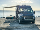 Relaxing RV rental scene: a black camper van by calm waters with an extended awning, table, chairs, and rug.