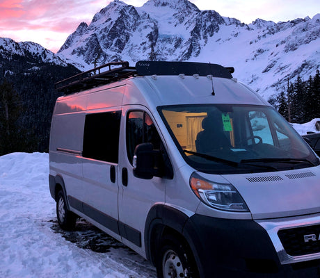 A silver RV rental is parked in a snowy landscape with majestic, snow-capped mountains and a vibrant pink sunrise sky.