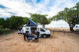 Two travelers high-fiving by their RV rental camper van with rooftop tent, parked on a sandy beach near the ocean.
