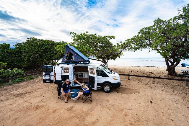 Two travelers high-fiving by their RV rental camper van with rooftop tent, parked on a sandy beach near the ocean.