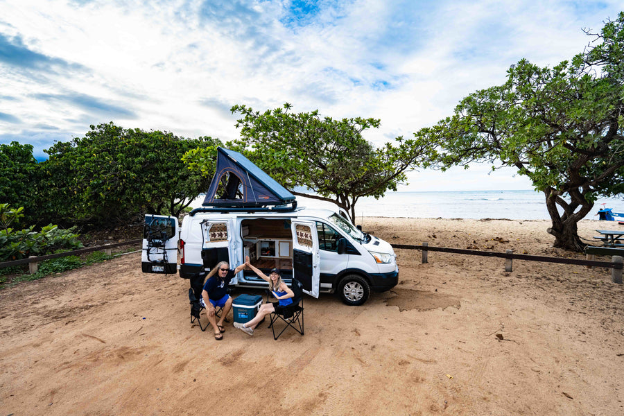 Two travelers high-fiving by their RV rental camper van with rooftop tent, parked on a sandy beach near the ocean.