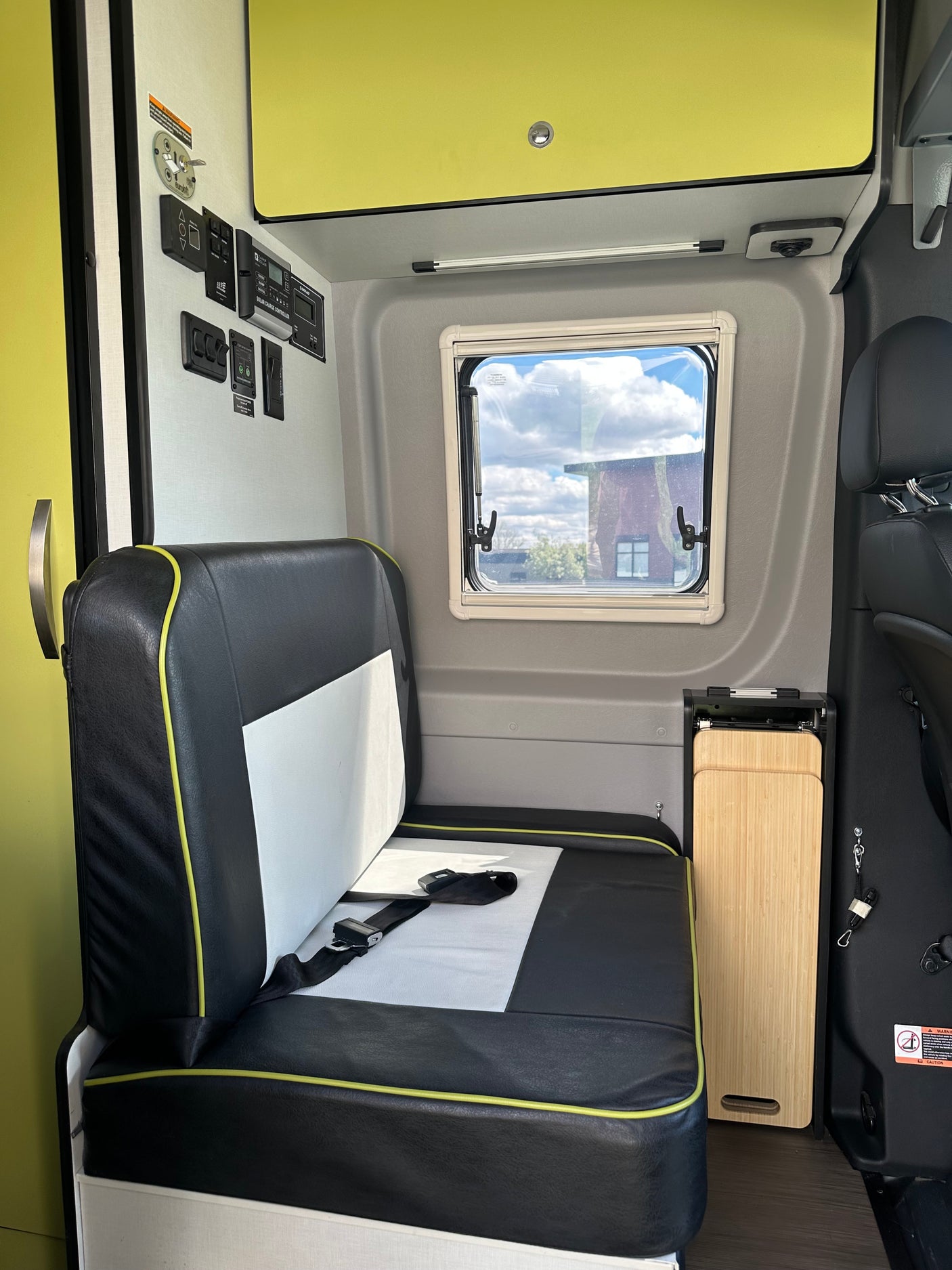View inside an RV rental showing a black/white seat with seatbelt, window, controls, and a folded wooden table.