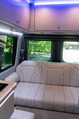 Modern RV rental interior with light-colored bench seating, bright purple overhead lights, and scenic views through the windows.