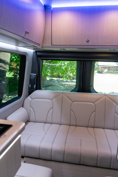 Modern RV rental interior with light-colored bench seating, bright purple overhead lights, and scenic views through the windows.