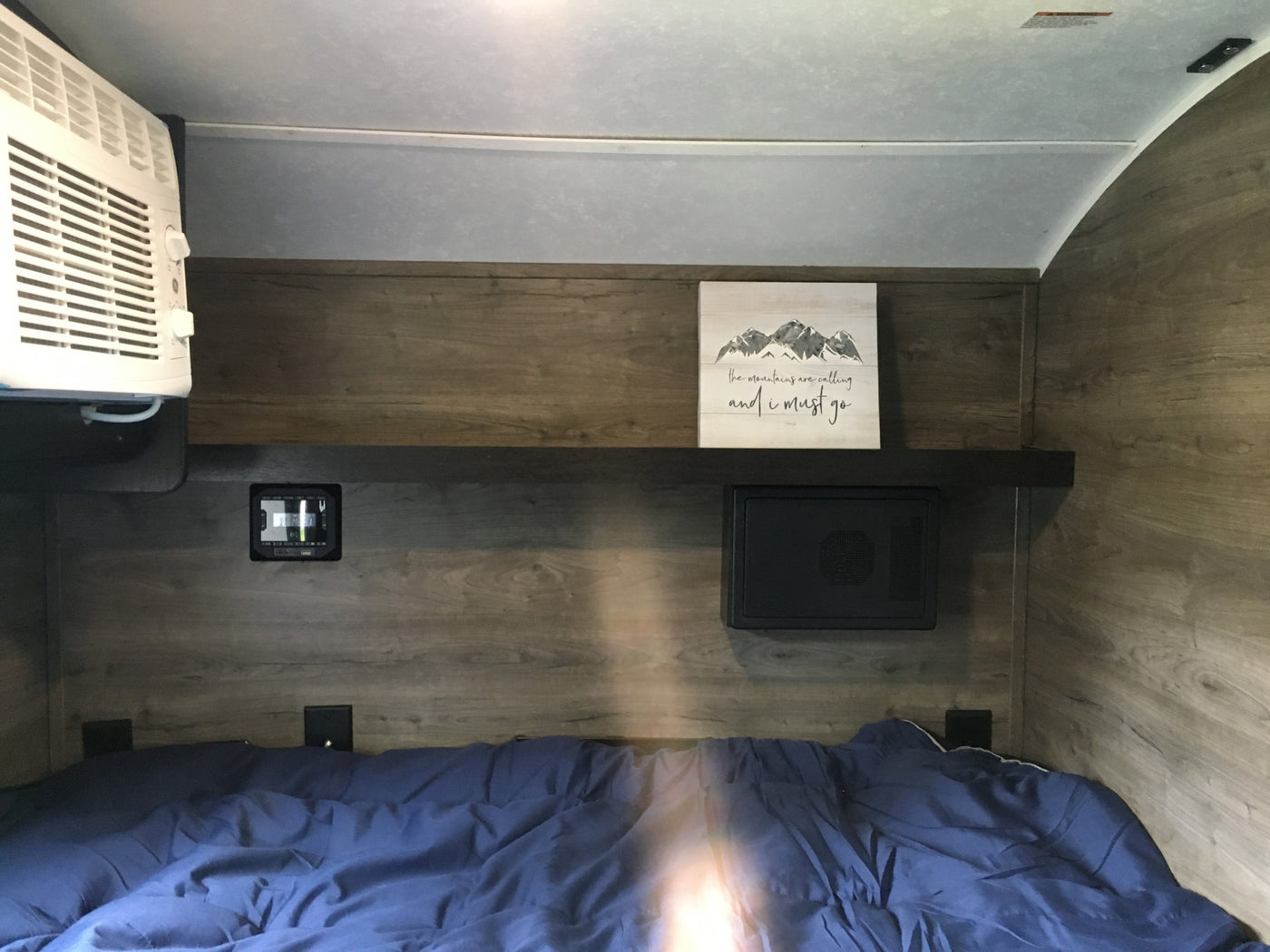 Cozy RV rental bedroom featuring a bed with blue covers, wall-mounted AC, electronic controls, and a shelf with mountain decor. — photo 9