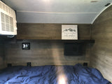 Cozy RV rental bedroom featuring a bed with blue covers, wall-mounted AC, electronic controls, and a shelf with mountain decor.