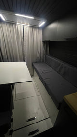 Cozy RV rental interior featuring a bright white table, grey bench seating, curtains, and LED lighting overhead.