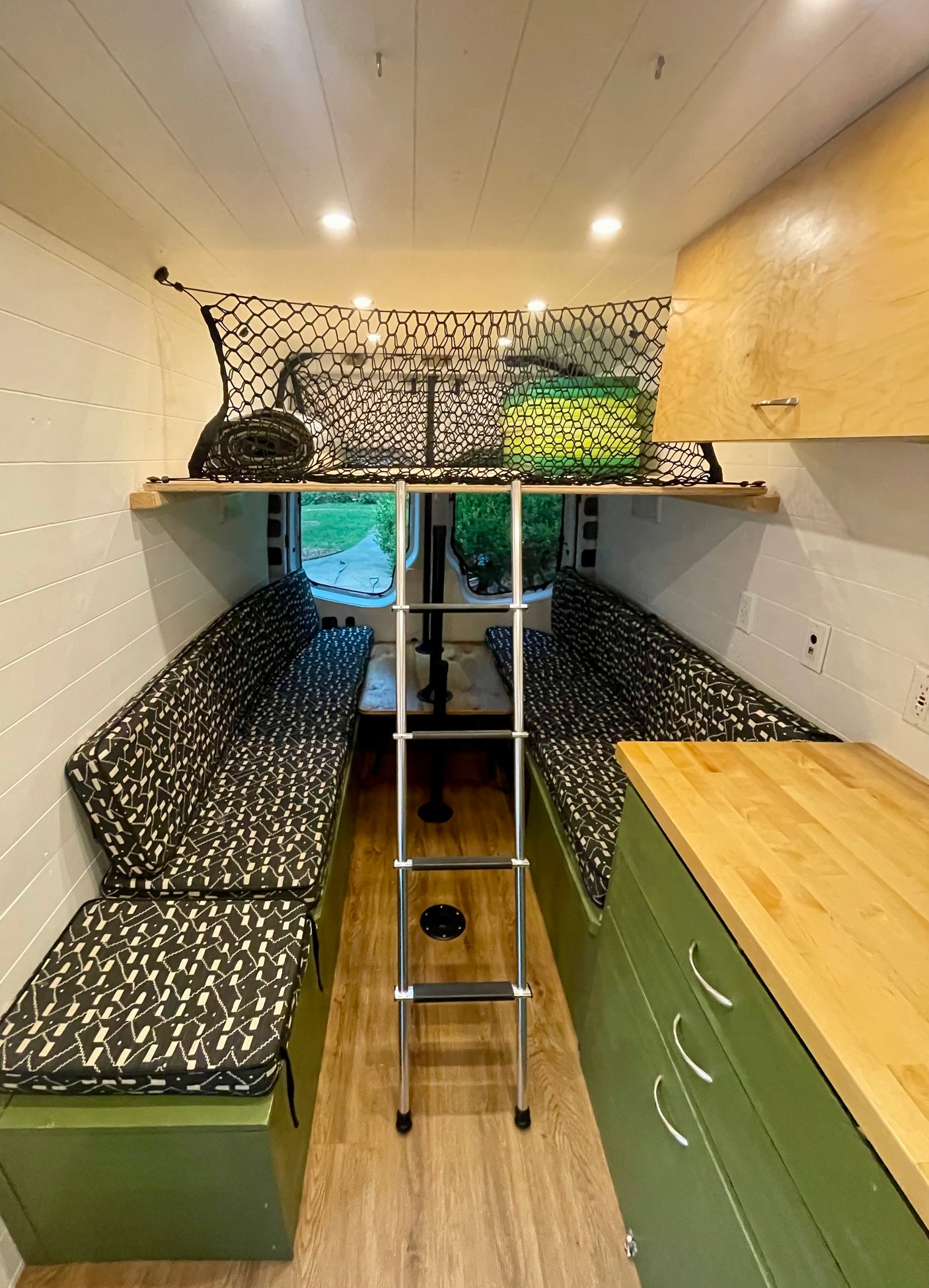 This rv rental's organized camper van interior features patterned seating, a ladder, overhead storage nets, and a compact kitchen.