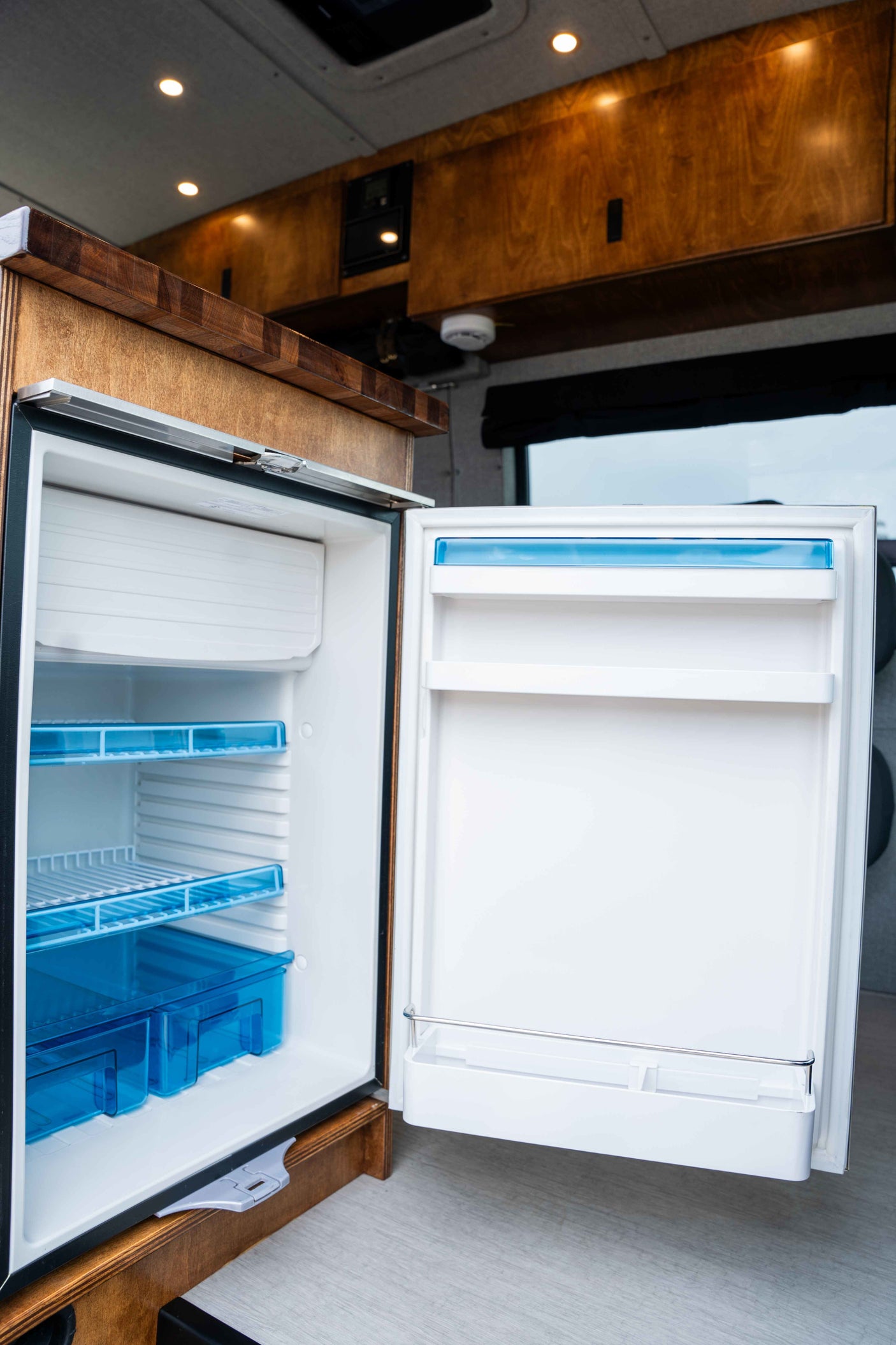 This RV rental features a clean, open refrigerator with blue shelving, nestled within a stylish wooden interior for your adventures.