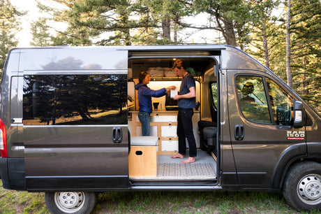 Inside a custom rv rental camper van, a couple stands by the kitchen. Ideal for van life adventures among the trees.
