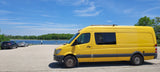 This vibrant yellow RV rental van is perfectly parked by a tranquil lake, surrounded by lush trees under a clear blue sky.