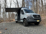 Explore off-road with this grey Sprinter van RV rental, ready for adventure with its awning extended in the woods.
