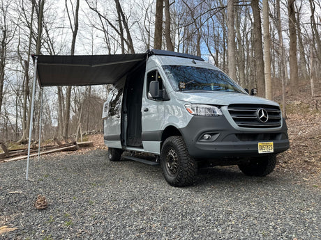 Explore off-road with this grey Sprinter van RV rental, ready for adventure with its awning extended in the woods.