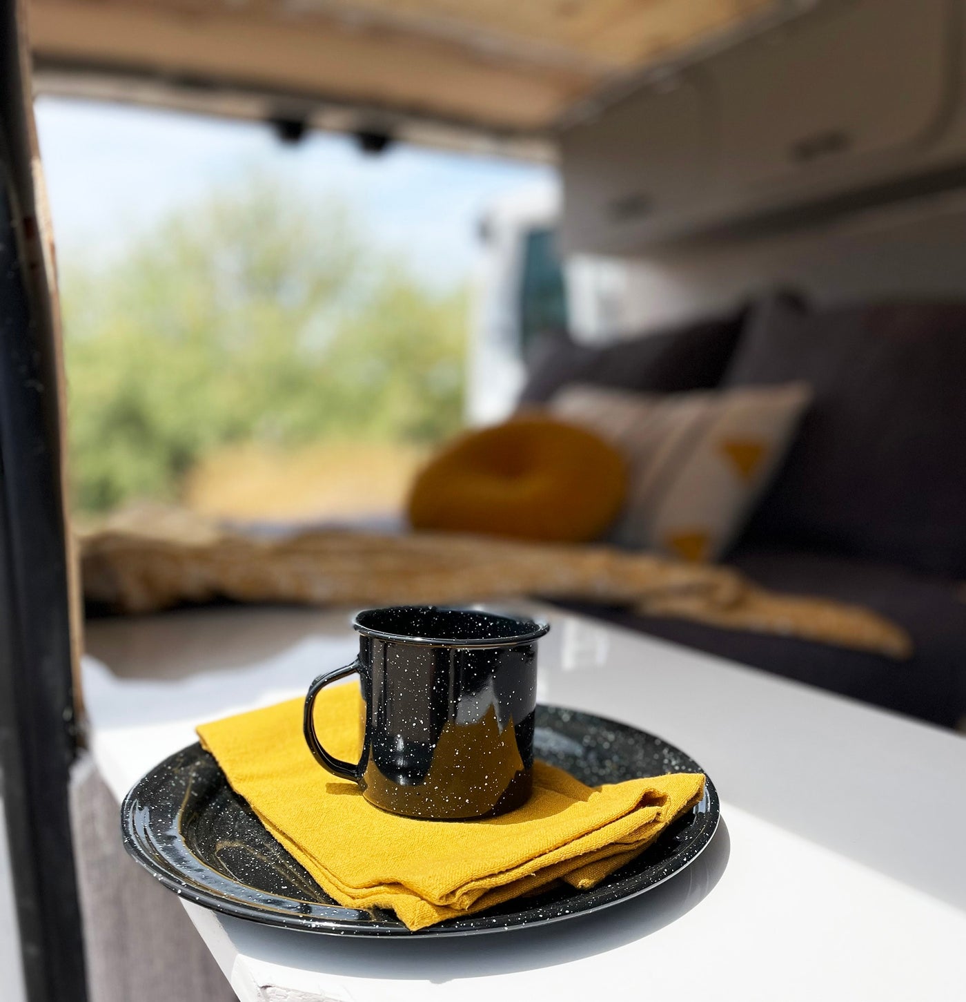 Cozy RV rental interior setup: black speckled mug, plate, and yellow napkin on a white table, with a blurred natural view. — photo 11