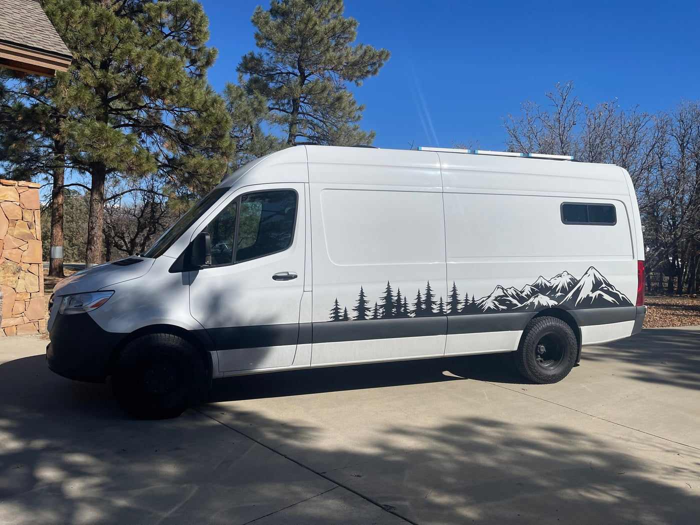 White camper van featuring tree and mountain graphics on a sunny driveway, ideal for an RV rental. — photo 8