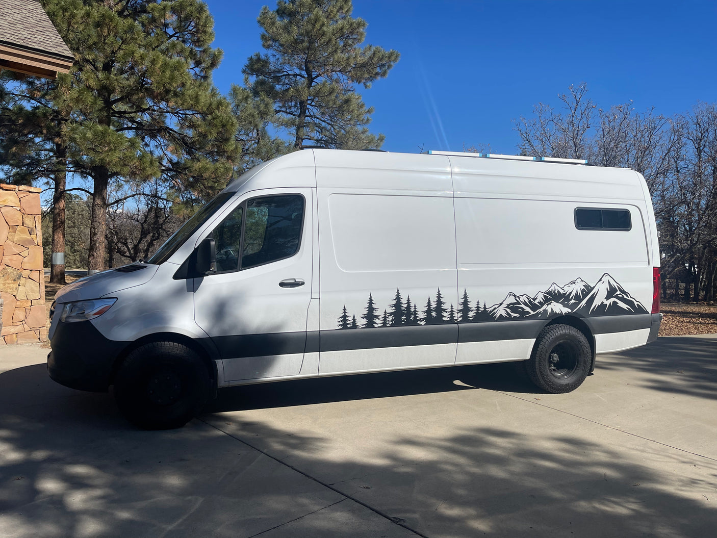 White camper van featuring tree and mountain graphics on a sunny driveway, ideal for an RV rental.