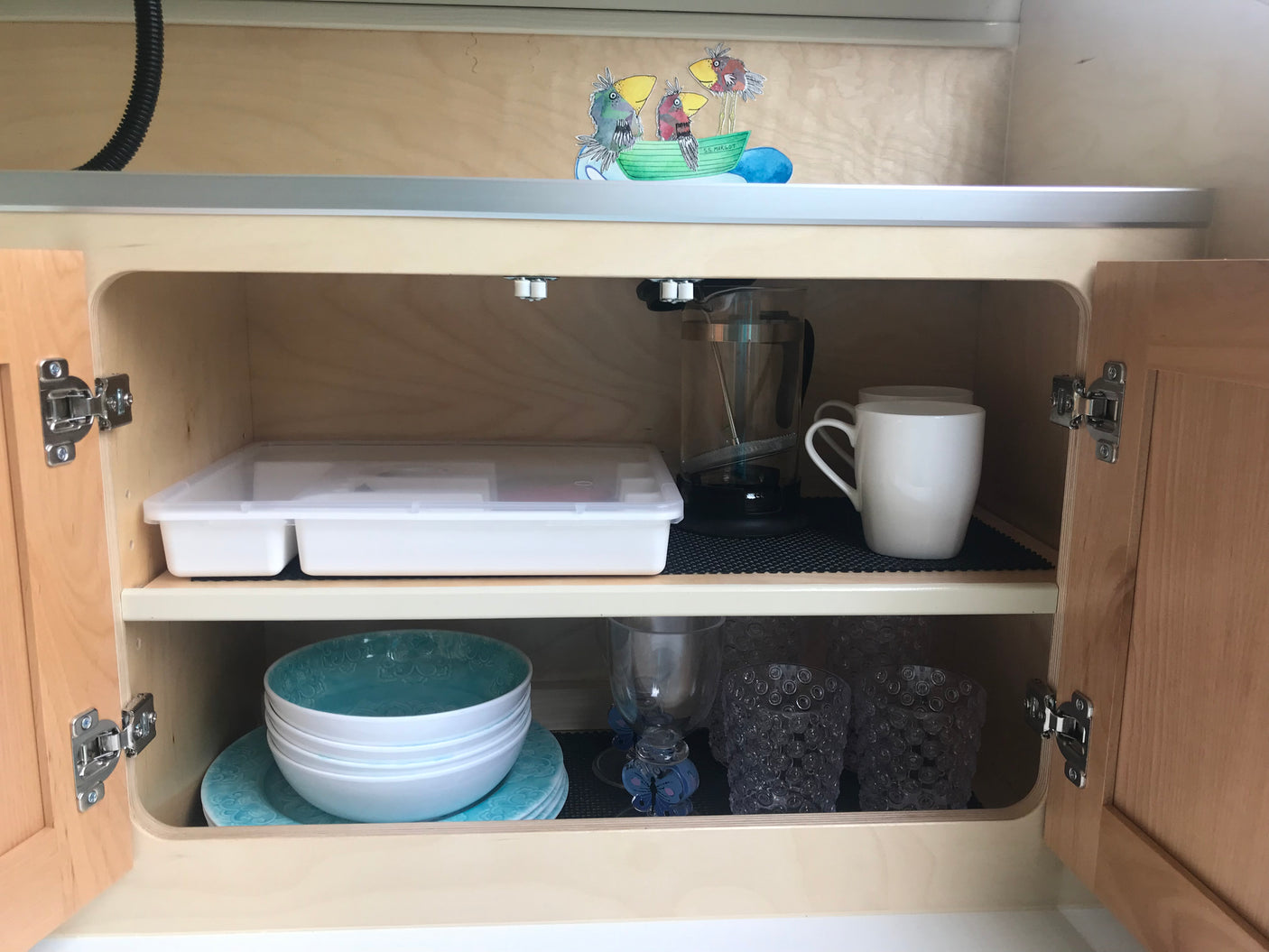 RV rental kitchen cabinet: Organized French press, mugs, bowls, plates, glasses, storage, and decorative birds in a boat.