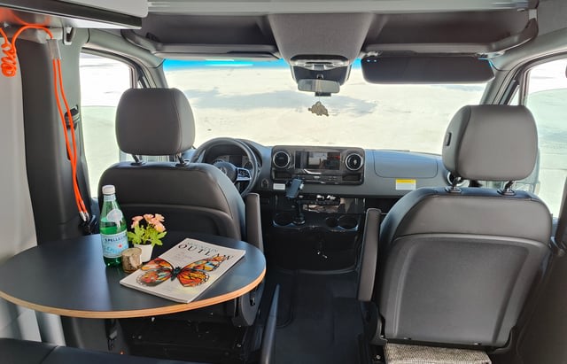 View into an rv rental camper van from the rear. Features a small round table, water, and magazine, with swivel front seats.