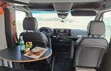 View into an rv rental camper van from the rear. Features a small round table, water, and magazine, with swivel front seats.