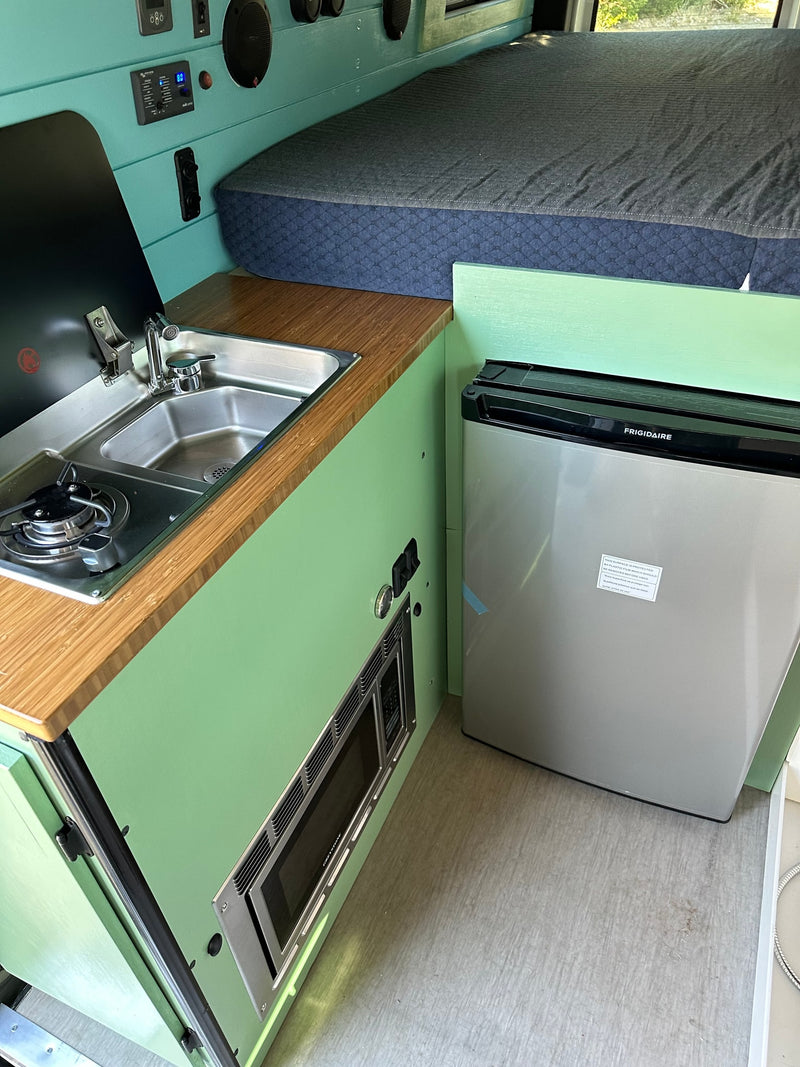 RV rental interior showcasing a modern kitchen with stove, sink, and microwave, plus a compact fridge and cozy bed. — photo 10