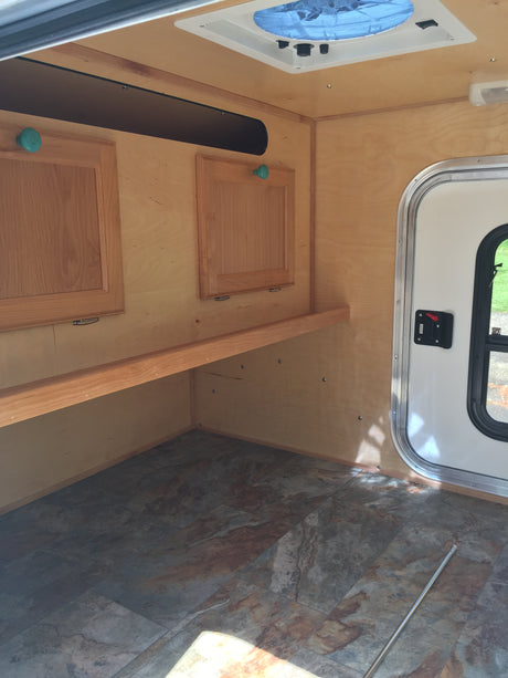 RV rental interior: Bright wood walls, patterned floor, overhead fan, window, and wooden storage compartments.