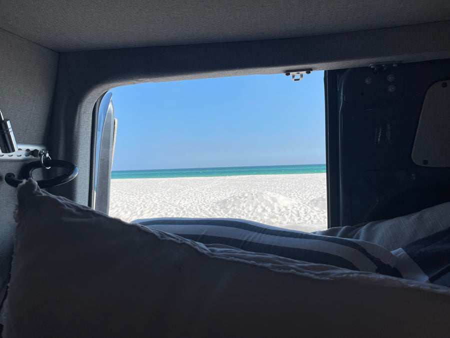 Wake up to paradise! RV rental bedroom view of white sand beach & clear turquoise ocean. Perfect campervan escape.