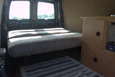 Cozy RV rental van interior: bed, wood storage, bright windows, and a rug. Perfect for your next adventure! — photo 9