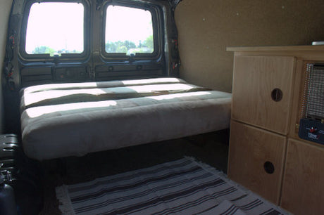 Cozy RV rental van interior: bed, wood storage, bright windows, and a rug. Perfect for your next adventure!