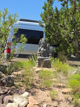A silver RV rental is parked behind a stone statue and arid plants, creating a tranquil outdoor scene under a clear blue sky.