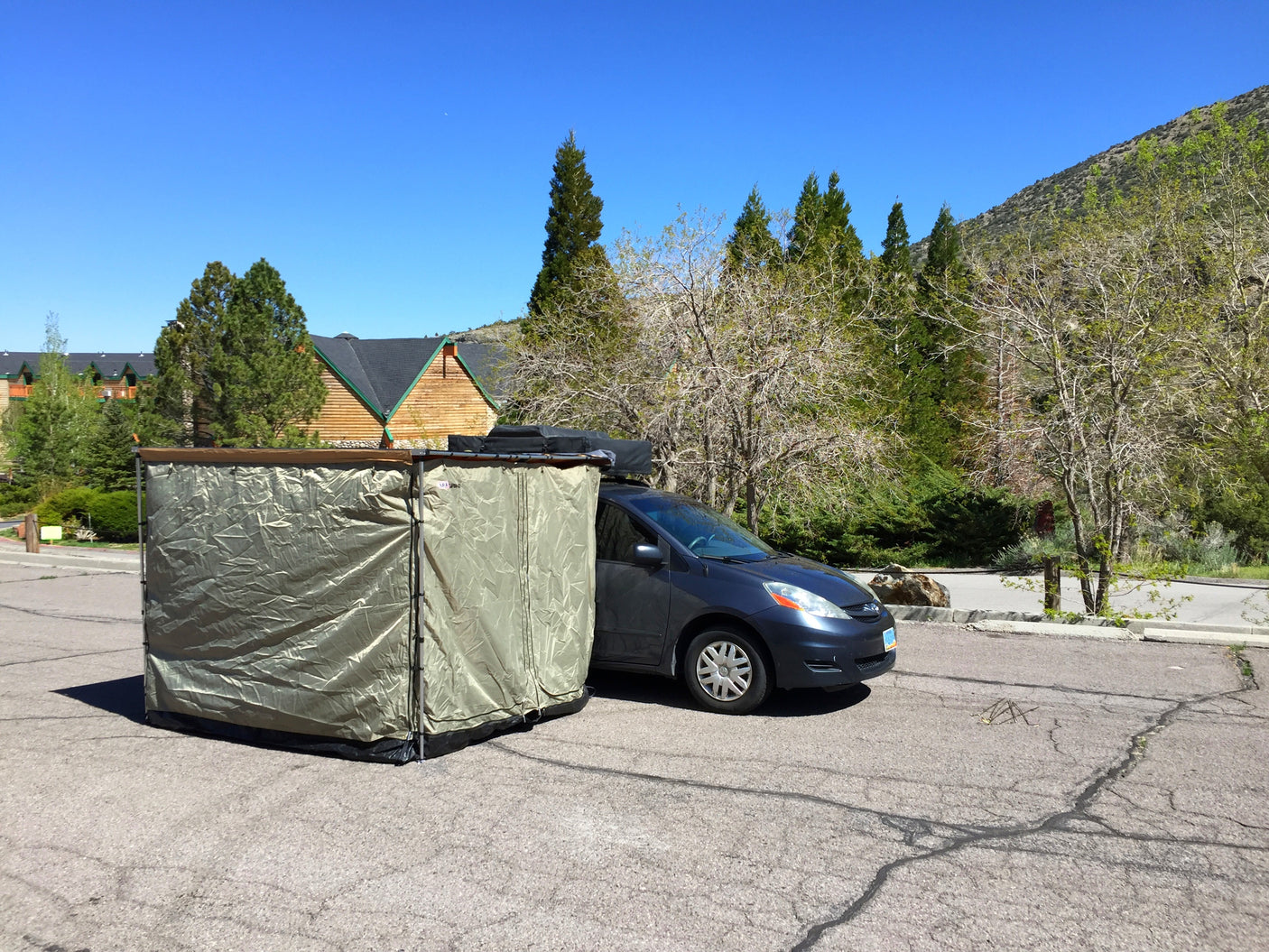 This RV rental setup features a dark blue minivan with an attached privacy tent, parked outdoors near a lodge.