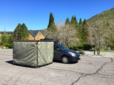 This RV rental setup features a dark blue minivan with an attached privacy tent, parked outdoors near a lodge.