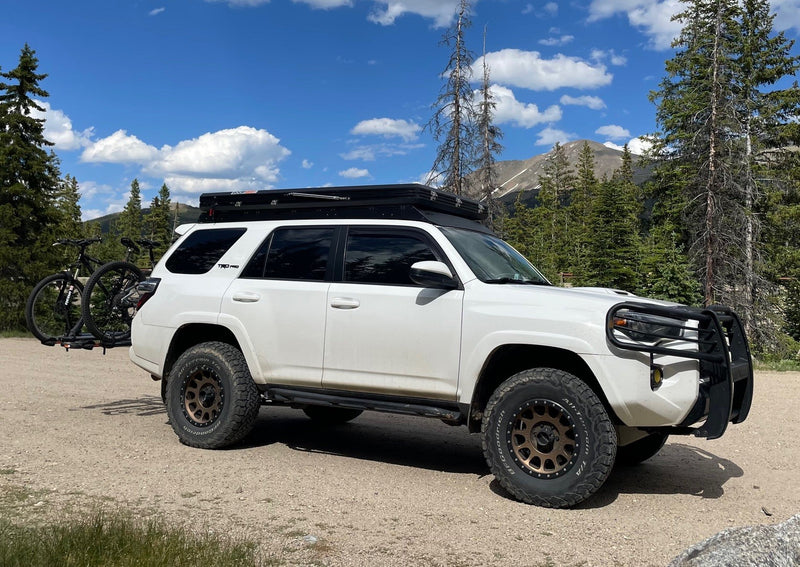 White 4Runner with rooftop tent, bike rack, & mountain tires on a dirt road by trees & mountains. Great for RV rental adventures. — photo 14