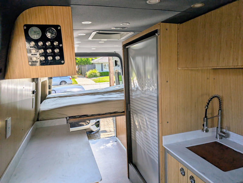 Clean RV rental interior featuring a compact kitchen, comfortable bed, control panel, and wood finishes for adventure travel. — photo 6