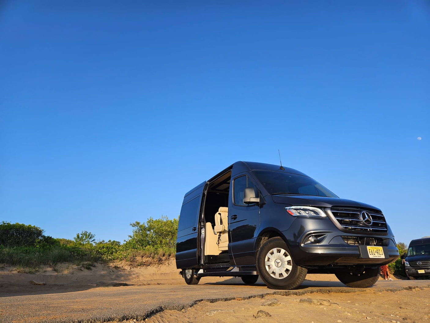 Dark gray Mercedes-Benz Sprinter van for RV rental, parked on a dirt road. Side door open. Clear blue sky with a faint moon.