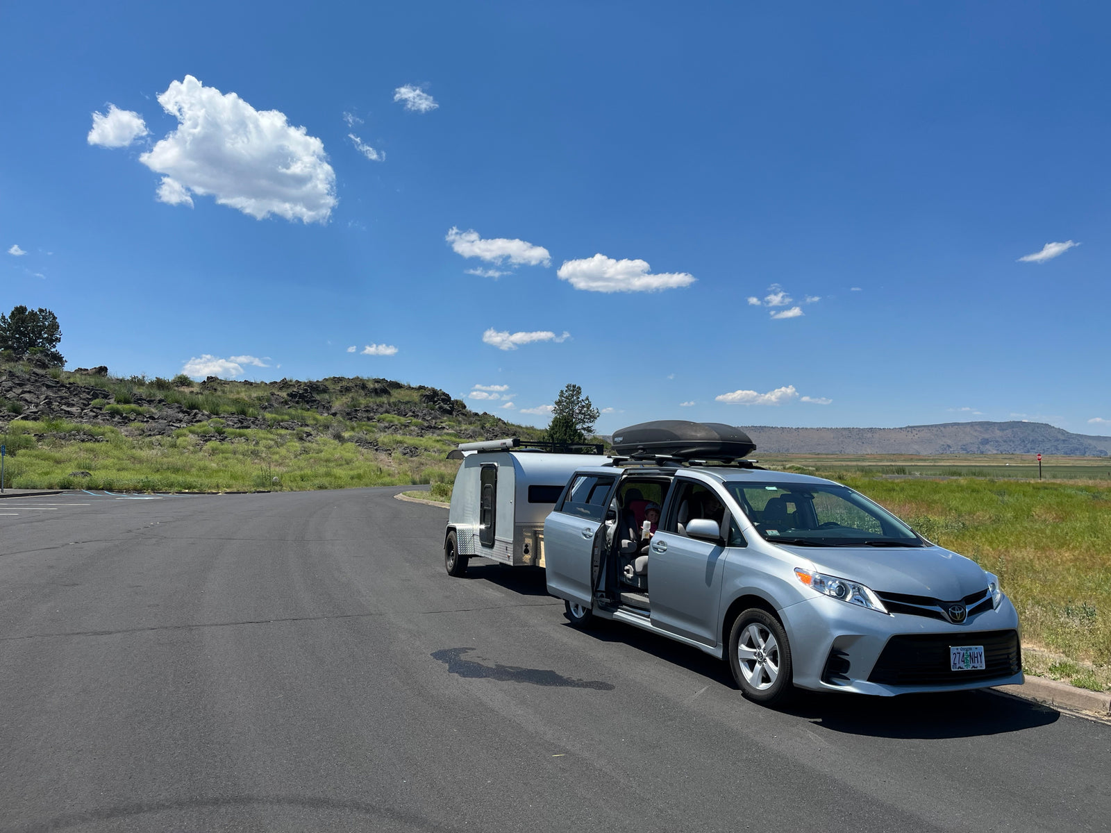 Silver minivan with an RV rental teardrop trailer on a sunny road, a child visible inside. Scenic rocky landscape in background.
