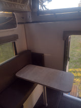 Inside an RV rental: a comfortable dinette with a light-colored table and dark cushioned seating, plus windows overlooking nature.