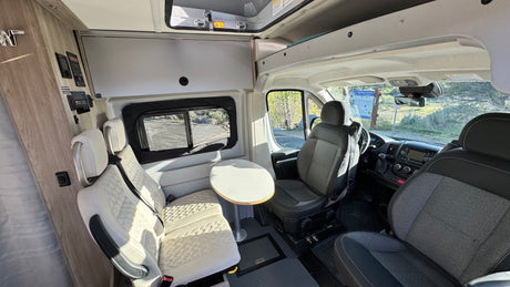 Cozy RV rental interior: Swiveling front seats, a dining area with light passenger seats, and control panels visible.