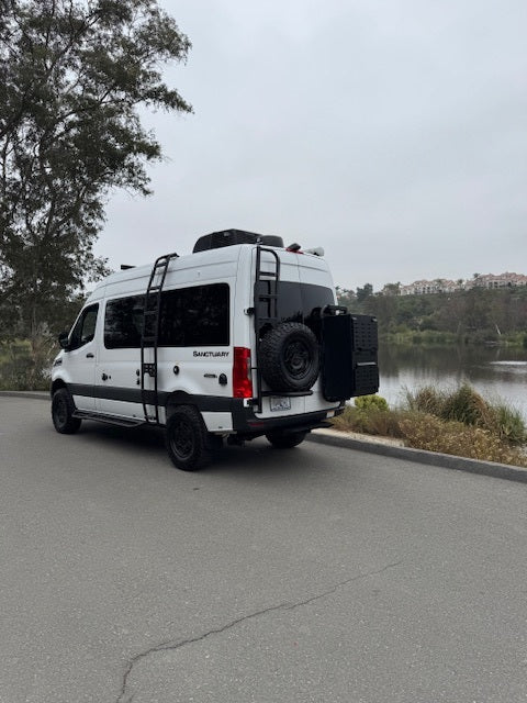 This white camper van RV rental, equipped with ladders and a spare tire, is parked by a lake, ready for adventure travel.
