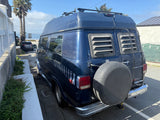 Blue RV rental van with a high top, rear ladder, and roof rack. Parked on a street overlooking the sunny ocean.