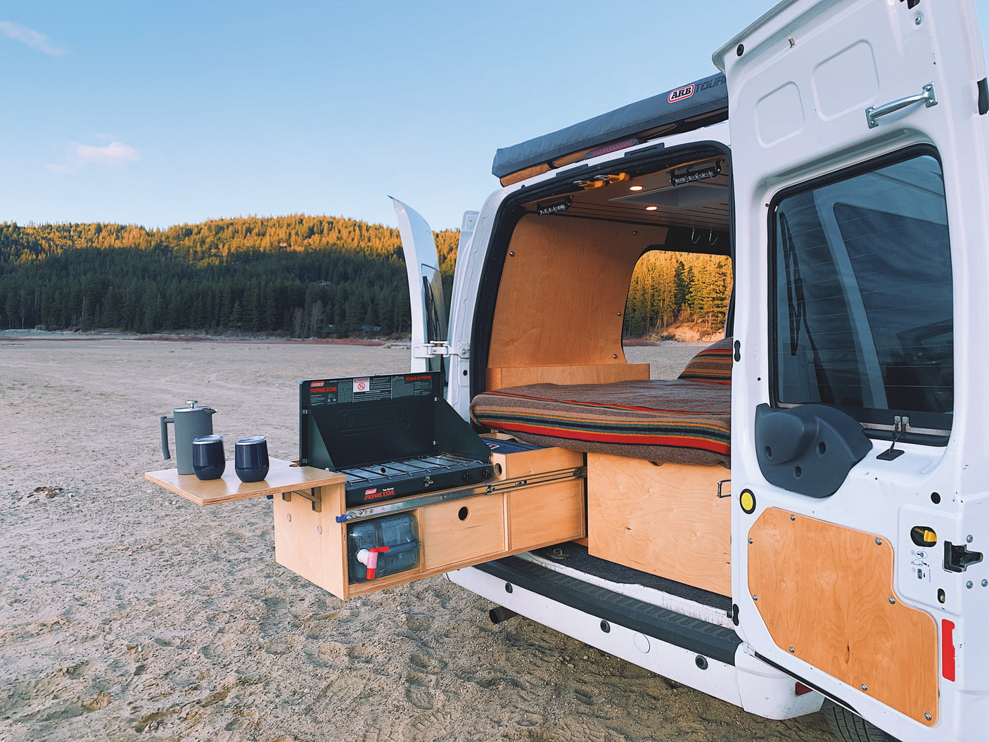An RV rental camper van with a cozy bed and outdoor kitchen setup, parked on a dry lakebed near a forest.