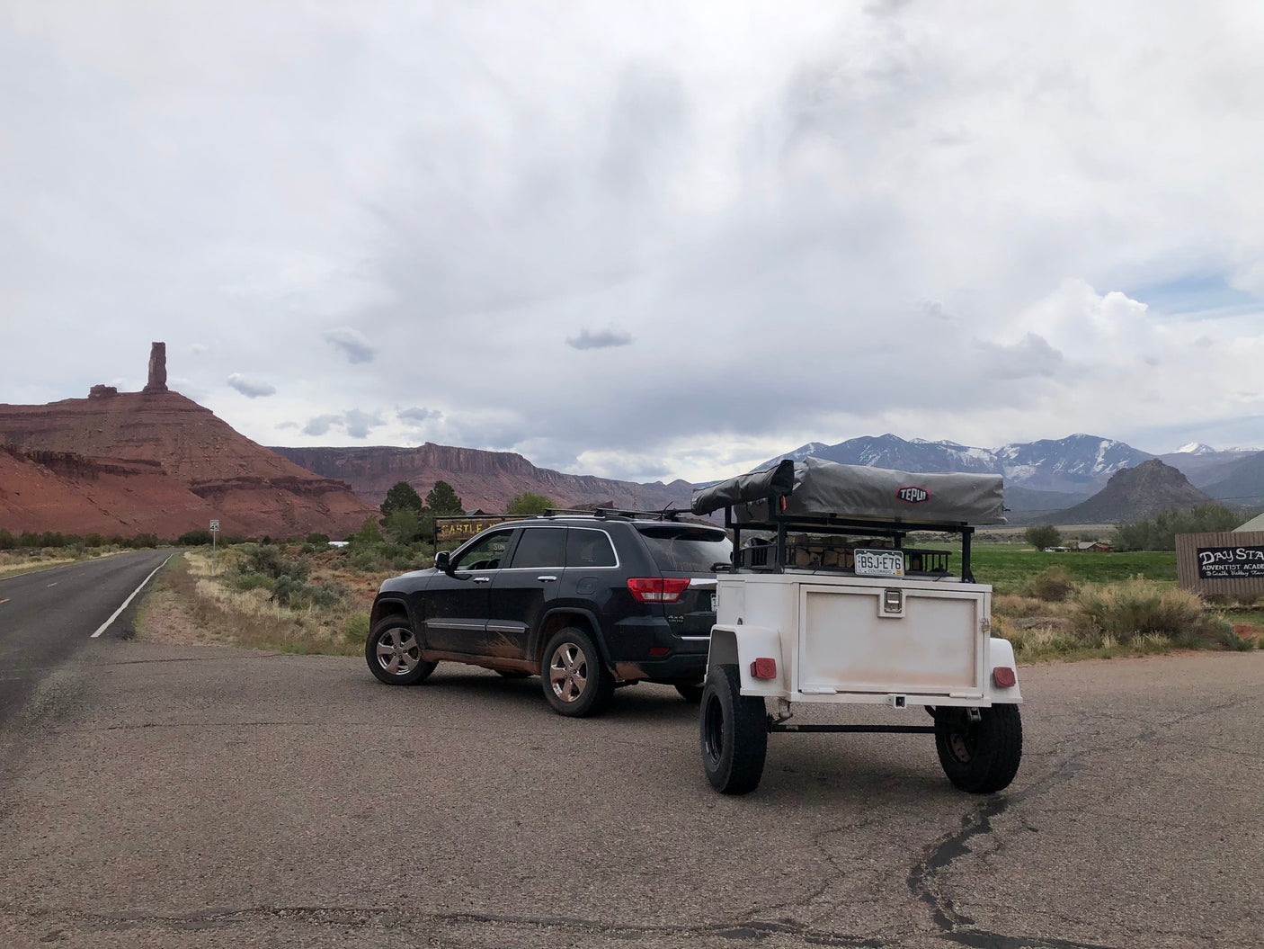 SUV towing RV rental trailer with rooftop tent amidst red rock formations and snowy mountains.