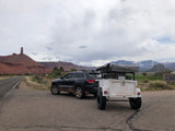 SUV towing RV rental trailer with rooftop tent amidst red rock formations and snowy mountains.