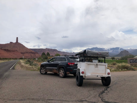 SUV towing RV rental trailer with rooftop tent amidst red rock formations and snowy mountains.
