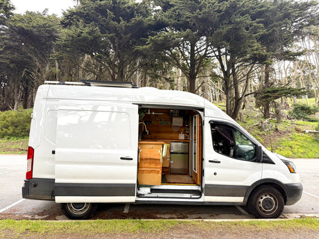Experience van life! This white rv rental campervan, open to show its wooden interior and kitchen, is parked by lush cypress trees.