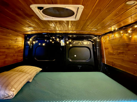 This cozy RV rental features a wooden interior, ceiling fan, warm string lights, and a comfy bed.