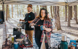 Outdoor cooking & relaxation with your RV rental. A couple prepares food and drinks under a canopy tent in the woods.