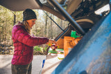 An RV rental provides kitchen convenience. A woman preps a red onion for cooking from the open trunk, surrounded by trees.
