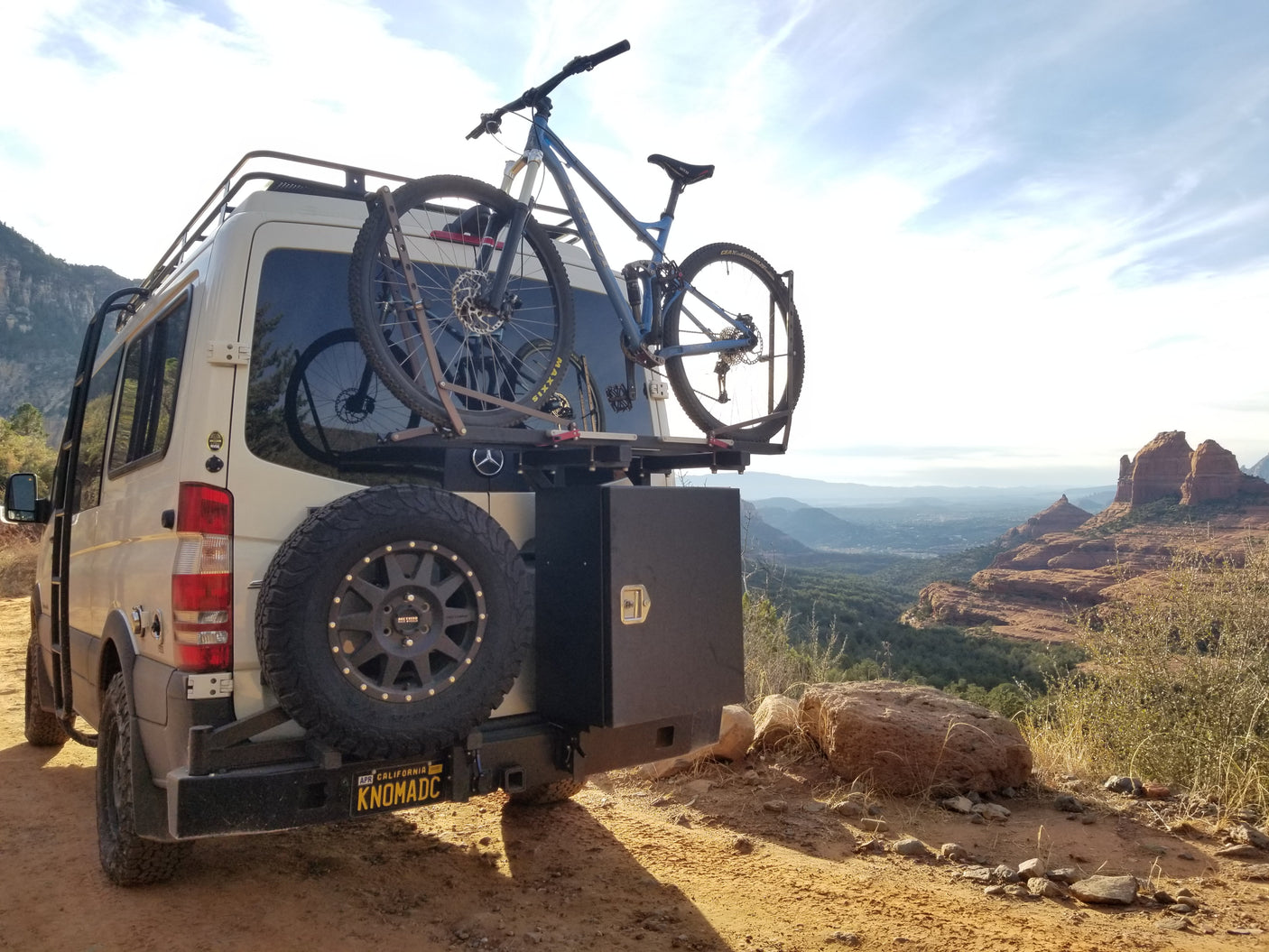 White Mercedes Sprinter rv rental van with a mountain bike & spare tire, parked at a breathtaking red rock overlook.